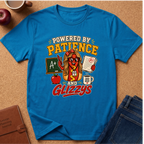 A bright blue graphic t-shirt laid flat on a cork background, featuring a cartoon hot dog character with glasses and the text 'POWERED BY PATIENCE AND GLIZZYS' along with school-themed icons like an apple, chalkboard, and coffee mug. Styled flat-lay props include a notebook, apple, coffee cup, and jeans.
