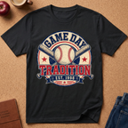 A black  short-sleeve graphic T-shirt laid flat on a tan cork-like surface, featuring a baseball-themed circular print with the words “GAME DAY TRADITION EST. 1998,” crossed bats, a baseball, and stars.