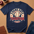 A navy blue short-sleeve graphic T-shirt laid flat on a tan cork-like surface, featuring a baseball-themed circular print with the words “GAME DAY TRADITION EST. 1998,” crossed bats, a baseball, and stars.