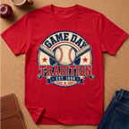 A red short-sleeve graphic T-shirt laid flat on a cork surface, featuring a baseball-themed circular print with the words 'GAME DAY TRADITION' and 'EST. 1998' around crossed bats and a baseball.