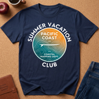 A navy blue graphic t-shirt laid flat on a brown cork board background, surrounded by lifestyle props including an apple, a coffee mug, a leather journal, and the corner of a pair of jeans. The t-shirt features a circular graphic with a topographic map design in teal and orange, with white text reading 'SUMMER VACATION PACIFIC COAST CLUB' and 'COASTAL MAPPING DEPT.