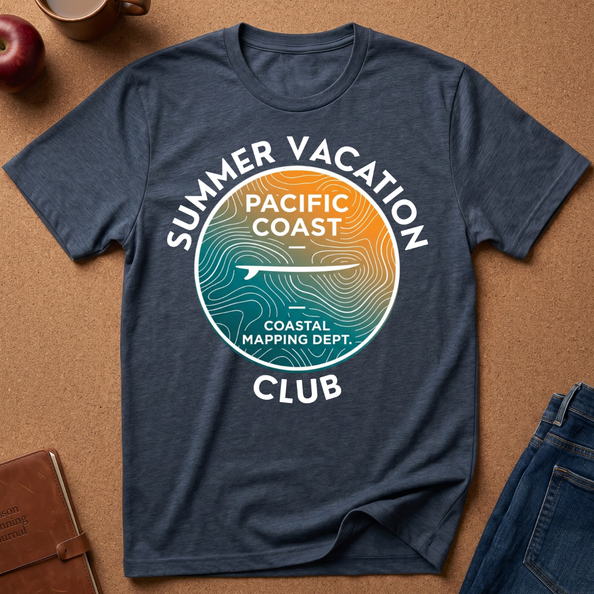 A navy blue heather graphic t-shirt laid flat on a brown cork background, featuring a circular 'Summer Vacation Pacific Coast Coastal Mapping Dept.' design in teal, orange, and white.