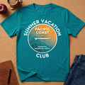 A teal short-sleeve graphic T-shirt laid flat on a cork surface, with a large circular coastal-themed print reading “SUMMER VACATION PACIFIC COAST CLUB” in white. The print includes a topographic map design and airplane silhouette, with a warm orange-to-yellow gradient inside the circle.
