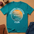 A teal short-sleeve graphic T-shirt laid flat on a cork surface, with a large circular coastal-themed print reading “SUMMER VACATION PACIFIC COAST CLUB” in white. The print includes a topographic map design and airplane silhouette, with a warm orange-to-yellow gradient inside the circle.