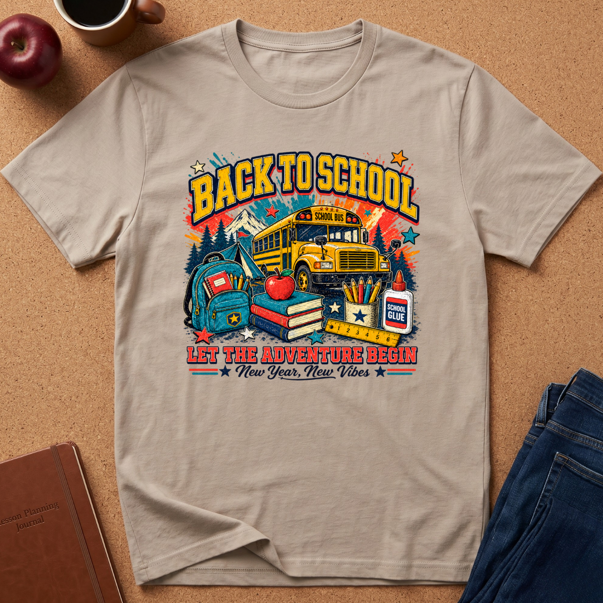 A beige graphic t-shirt laid flat on a brown corkboard background, surrounded by lifestyle props including an apple, a coffee cup, a notebook, and a pair of blue jeans. The t-shirt features a colorful 'Back to School' graphic with a school bus, books, and an apple.