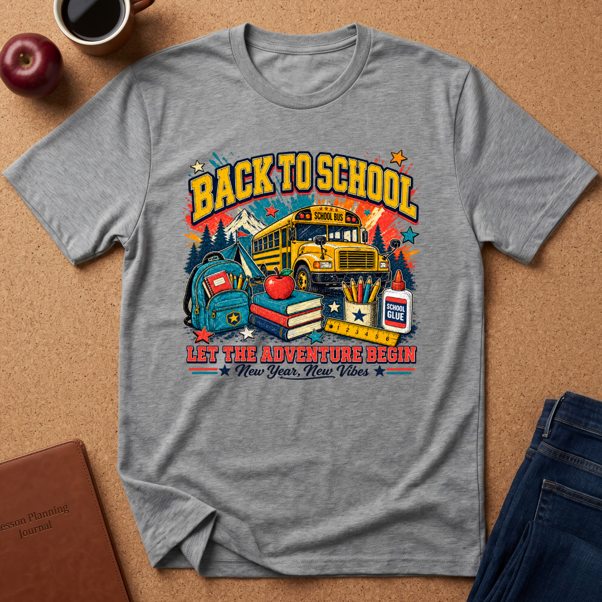 A heather gray graphic t-shirt laid flat on a cork background, featuring a colorful 'Back to School' design with a school bus, books, an apple, and the text 'Let the adventure begin New Year, New Vibe!'.