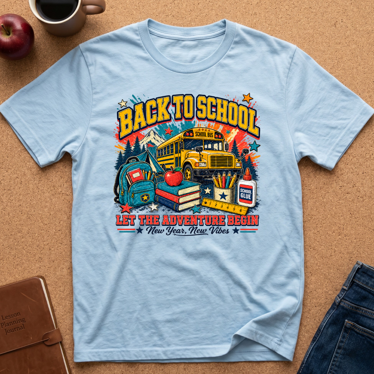 A light blue graphic t-shirt laid flat on a brown cork background, surrounded by lifestyle props including an apple, a coffee mug, a notebook, and blue jeans. The t-shirt features a colorful 'Back to School' graphic with a yellow school bus, books, an apple, and the text 'Let the adventure begin'.