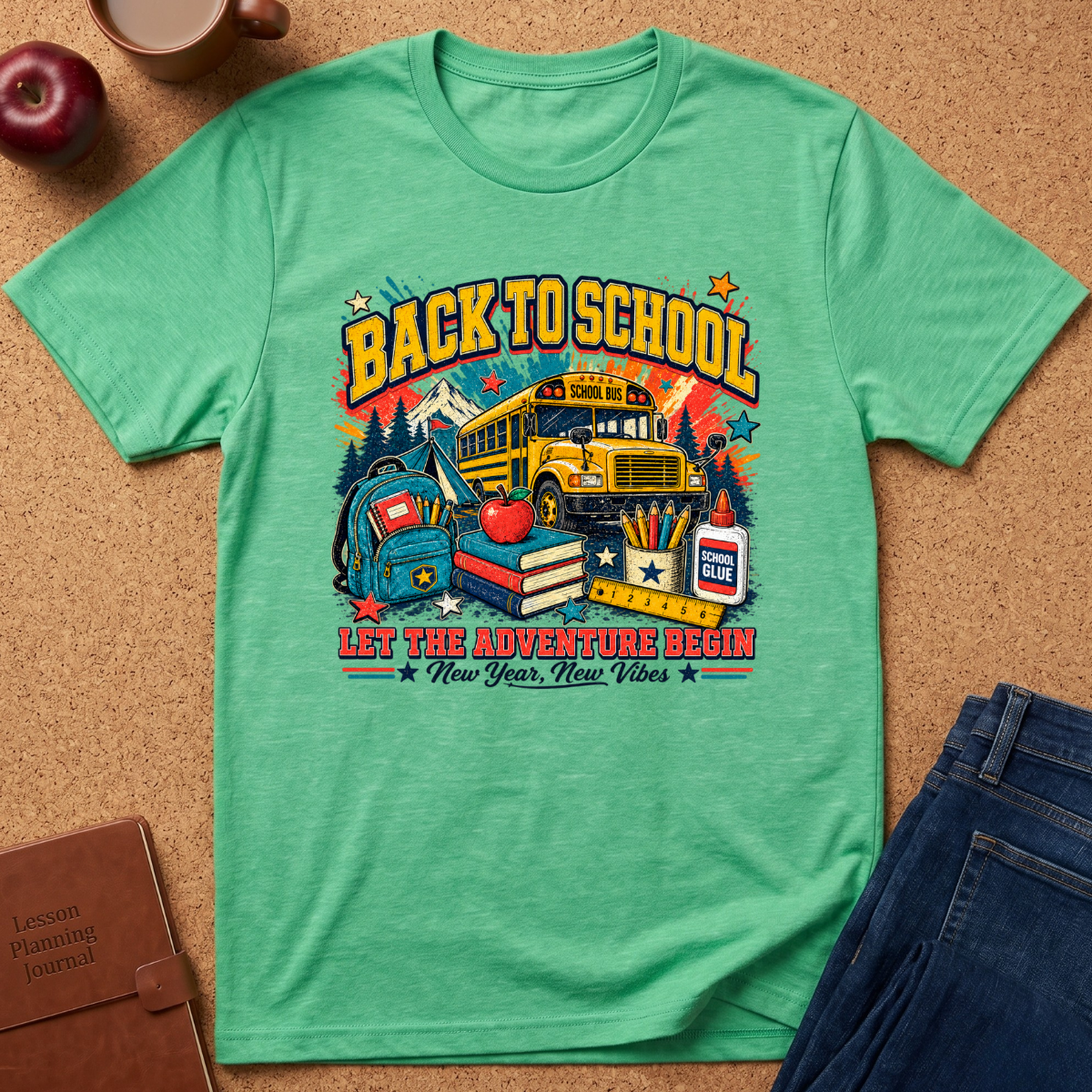 A mint green short-sleeve t-shirt with a colorful 'Back to School' graphic, laid flat on a cork background with props including an apple, coffee cup, book, and jeans.