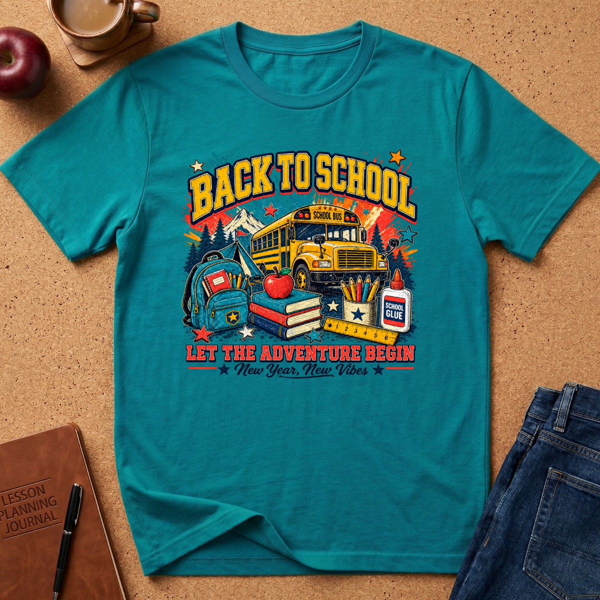 A teal short-sleeve graphic T-shirt laid flat on a cork surface, surrounded by school-themed props including a notebook, apple, coffee cup, and jeans. The shirt features a large retro-style 'Back to School' print with a school bus, books, apple, pencils, and stars.