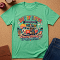 A mint green graphic t-shirt laid flat on a cork background, surrounded by lifestyle props including an apple, a coffee cup, a brown journal, and a pair of blue jeans. The t-shirt features a colorful retro-style illustration of a vintage VW bus loaded with school supplies and books, with the text 'OUT OF OFFICE EDUCATOR EDITION' and 'Creating Memories Not Lesson Plans'.