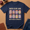 A navy blue graphic t-shirt laid flat on a brown cork background, surrounded by lifestyle props including an apple, a coffee cup, a notebook, and jeans. The t-shirt features a colorful graphic of eight labeled jars (Algebraic Equations, Calculus, Geometry, Ratios, Fractions, Problem Solving, Numbers, and Normals) with the text 'MATH IS MY JAM' and 'AND I'M HERE TO SPREAD IT!'.