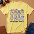 A yellow graphic t-shirt laid flat on a brown cork background, surrounded by lifestyle props including a coffee mug, an apple, a notebook, and blue jeans. The t-shirt features a colorful graphic of eight labeled jars (Algebraic Equations, Calculus, Geometry, Ratios, Fractions, Problem Solving, Numbers, Normals) with the text 'MATH IS MY JAM' above and 'AND I'M HERE TO SPREAD IT!' below.