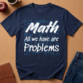A navy blue graphic t-shirt laid flat on a brown cork board background, surrounded by lifestyle props including a brown leather journal, a red apple, a coffee cup, and blue jeans. The t-shirt features a white graphic text reading 'Math All we have are Problems' over a light blue handwritten math equation background.