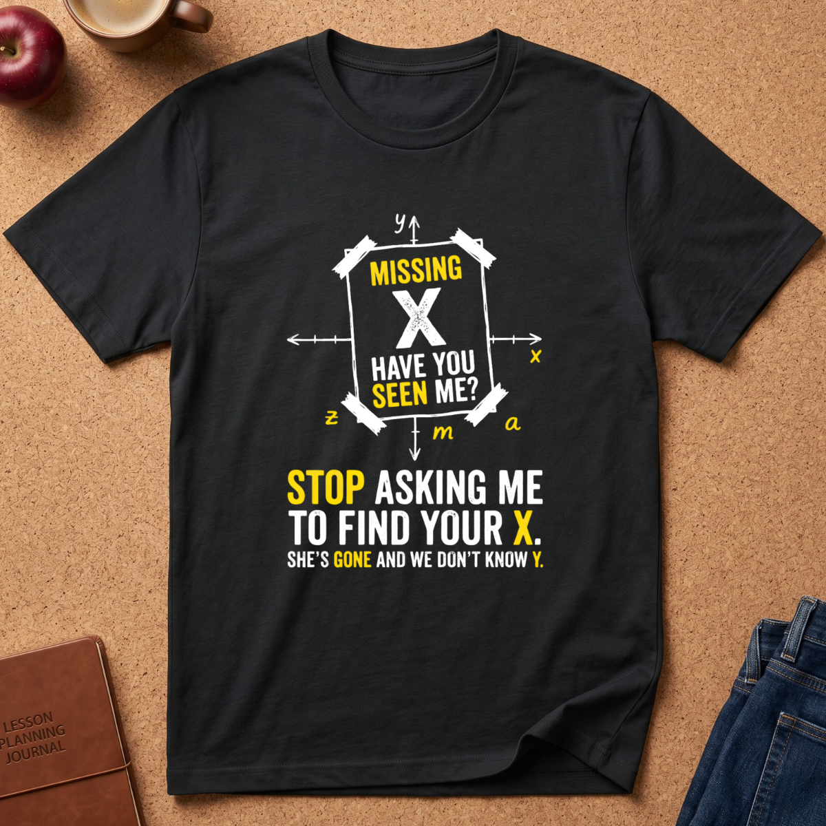 A black short-sleeve graphic T-shirt laid flat on a cork background, with a white and yellow text design about a missing X. Partial props around it include an apple, a mug, a journal, and jeans.