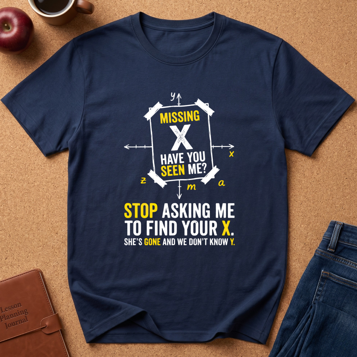 A navy blue graphic t-shirt laid flat on a brown cork board background, surrounded by lifestyle props including an apple, a coffee mug, a leather journal, and the corner of a pair of blue jeans. The t-shirt features a white and yellow graphic design with math equation symbols and the text 'MISSING X HAVE YOU SEEN ME? STOP ASKING ME TO FIND YOUR X.
