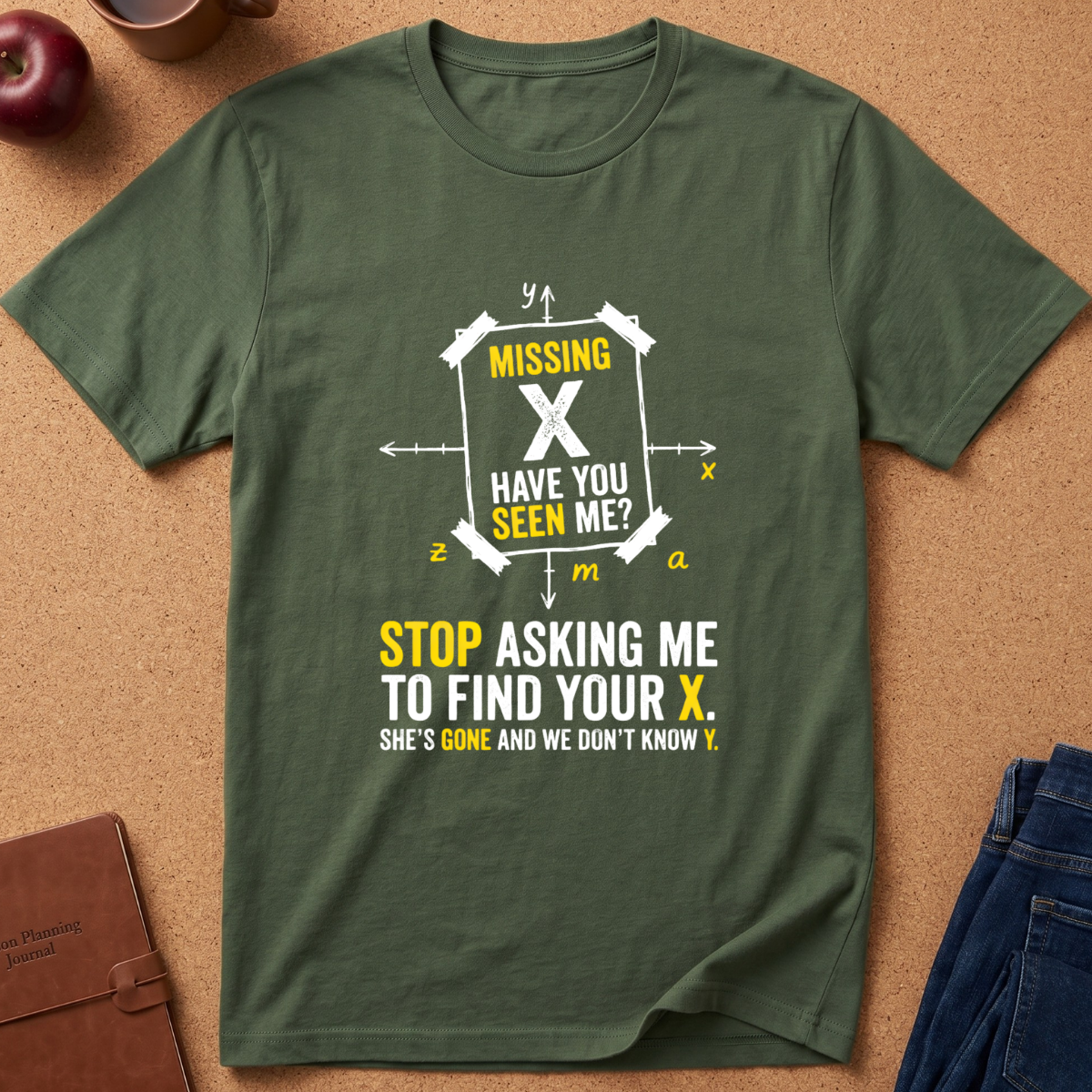 An olive green graphic t-shirt laid flat on a cork background, featuring a humorous 'missing X' message in white and yellow text with a geometric coordinate design.