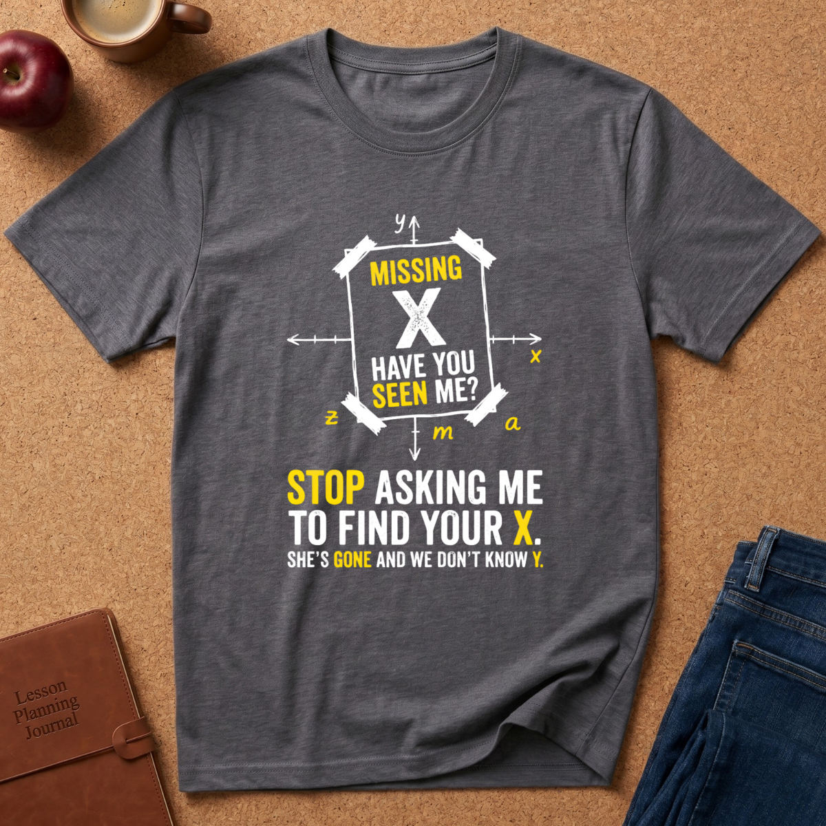 A dark heather gray graphic t-shirt laid flat on a brown cork surface, surrounded by lifestyle props including an apple, a coffee mug, a brown journal, and blue jeans. The t-shirt features a white and yellow graphic with math equation symbols and the text 'MISSING X HAVE YOU SEEN ME? STOP ASKING ME TO FIND YOUR X.
