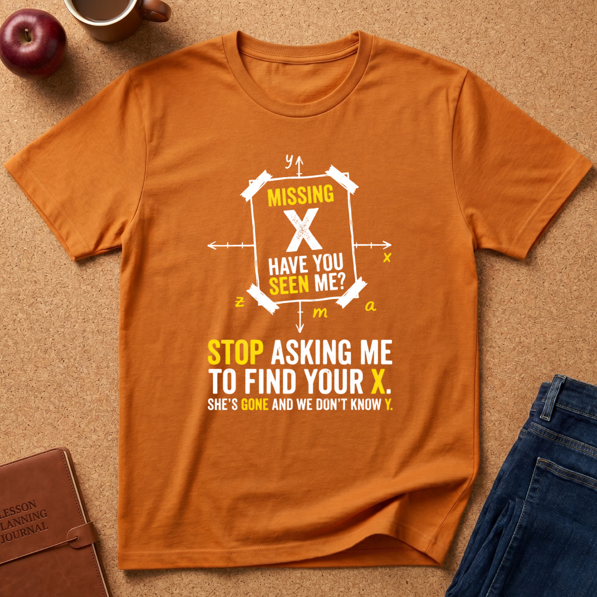 An orange short-sleeve graphic T-shirt laid flat on a cork surface, surrounded by lifestyle props including a brown leather journal, a red apple, a coffee mug, and blue jeans. The shirt features a white and yellow graphic with the text 'MISSING X HAVE YOU SEEN ME?' and 'STOP ASKING ME TO FIND YOUR X.