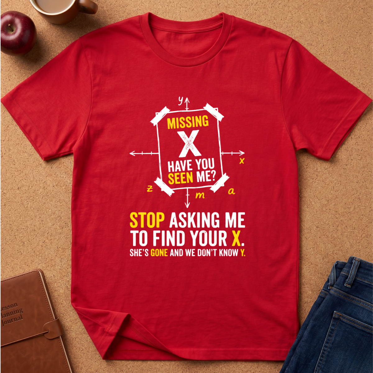 A red short-sleeve graphic T-shirt laid flat on a tan cork-like surface with styled props including an apple, a mug, a notebook, and blue jeans. The shirt has a large white and yellow text graphic reading 'MISSING X HAVE YOU SEEN ME? STOP ASKING ME TO FIND YOUR X.