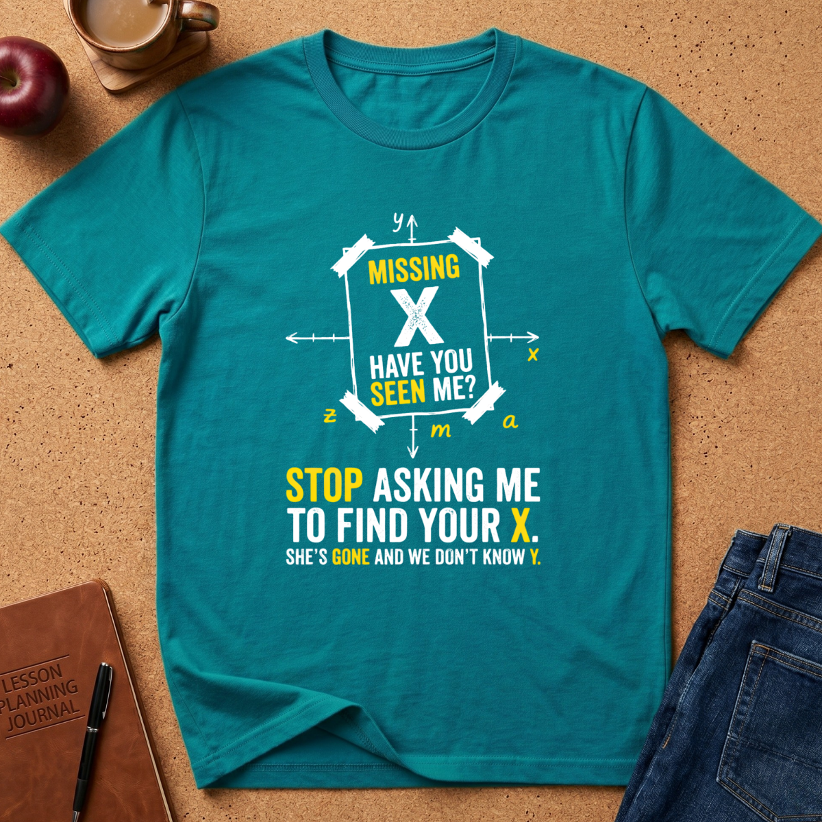 A teal short-sleeve graphic T-shirt laid flat on a cork surface, surrounded by lifestyle props including a coffee cup, apple, notebook, pen, and folded jeans. The shirt features a white and yellow printed design with the text 'MISSING X HAVE YOU SEEN ME? STOP ASKING ME TO FIND YOUR X.