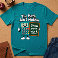 A teal graphic t-shirt laid flat on a cork surface, featuring a cartoon calculator character holding a chalkboard that reads 'Show your work' with the phrase 'The Math Ain't Mathin' above it. Styled flat-lay props include an apple, a coffee cup, a journal, a pen, and blue jeans.