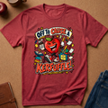 A heathered red short-sleeve t-shirt laid flat on a brown cork-like surface, featuring a colorful cartoon graphic of a smiling red apple surrounded by books, pencils, and papers with the text 'OFF TO CAUSE A KERFUFFLE!'. Partial jeans, a notebook, and a cup are visible around the shirt as styled props.
