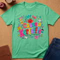 A mint green graphic t-shirt laid flat on a cork background, surrounded by lifestyle props including an apple, a coffee mug, a notebook, and a pair of blue jeans. The t-shirt features a colorful cartoon graphic with the text 'Off to Cause a Kerfuffle' in playful multicolored lettering.