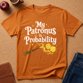 A orange graphic t-shirt laid flat on a brown cork board background, featuring the text 'My Patronus is Probability' in white with yellow stars and a wand-and-dice illustration. Surrounding props include an apple, a coffee cup, a brown journal, and folded blue jeans.