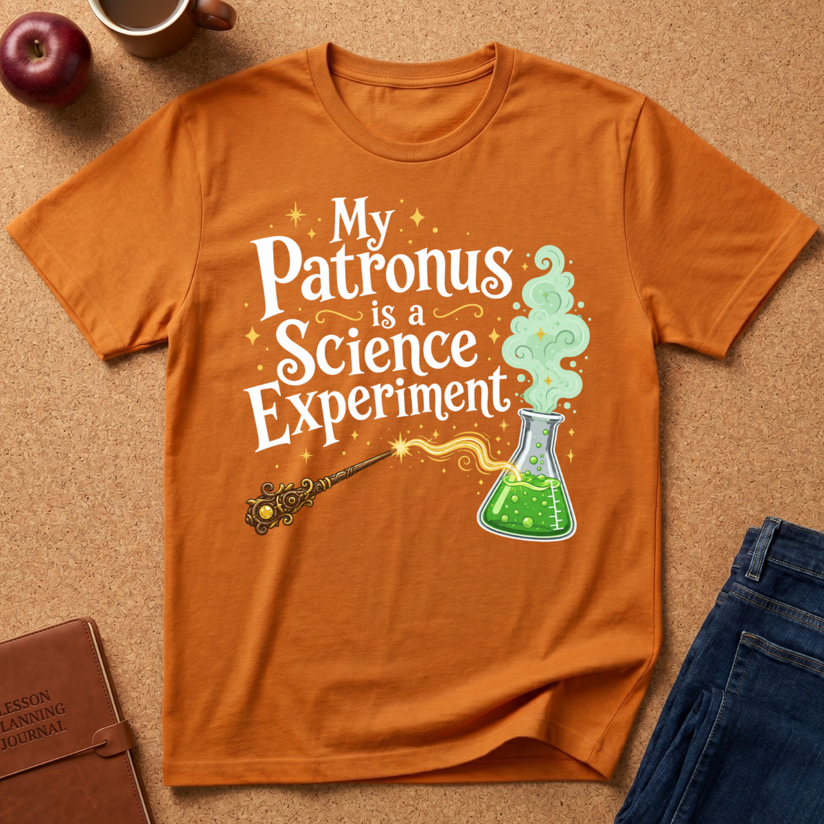 An orange graphic t-shirt laid flat on a cork background, featuring the text 'My Patronus is a Science Experiment' with a wand and green potion flask illustration. Styled flat-lay props include an apple, mug, notebook, and jeans.