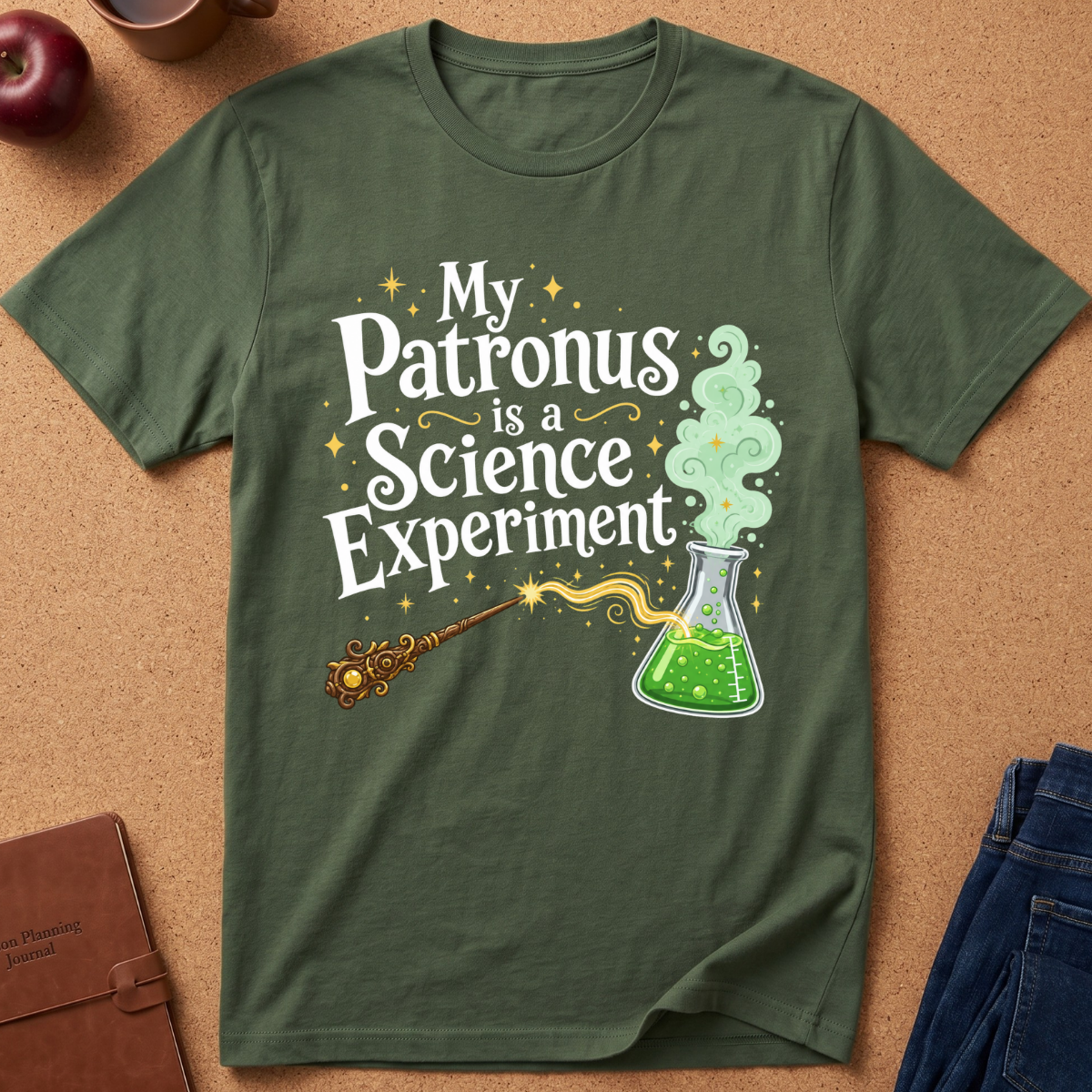 An olive green graphic t-shirt laid flat on a cork board background, featuring the text 'My Patronus is a Science Experiment' with a wand and a green potion flask illustration.