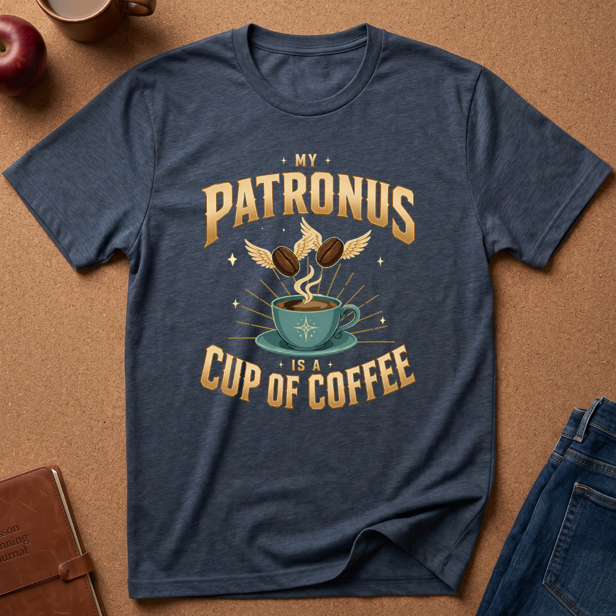 A navy blue graphic t-shirt laid flat on a brown cork background, surrounded by lifestyle props including an apple, a leather notebook, and folded blue jeans. The t-shirt features a retro-style gold and teal graphic with the text 'MY PATRONUS IS A CUP OF COFFEE' and an illustration of a teacup, coffee beans, and wings.