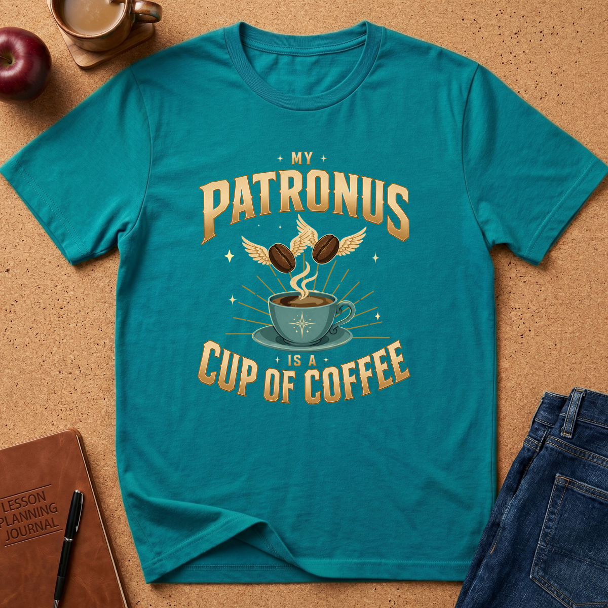 A teal graphic t-shirt laid flat on a cork surface, featuring a coffee-themed design with the text 'MY PATRONUS IS A CUP OF COFFEE' and an illustration of a steaming cup, coffee beans with wings, and stars. Styled flat-lay props include a notebook, pen, apple, mug, and jeans.