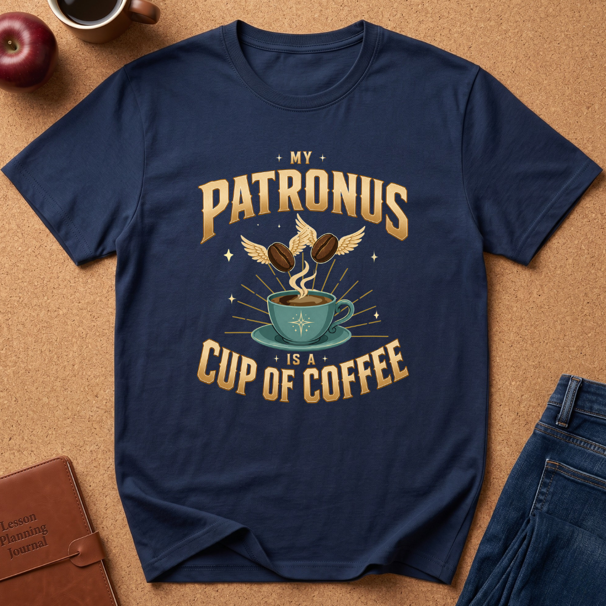 A navy blue graphic t-shirt laid flat on a cork background, featuring a vintage-style gold and teal print that reads 'MY PATRONUS IS A CUP OF COFFEE' with an illustration of a teacup, coffee beans, and a winged owl. Styled flat-lay props include an apple, a leather journal, and blue jeans.