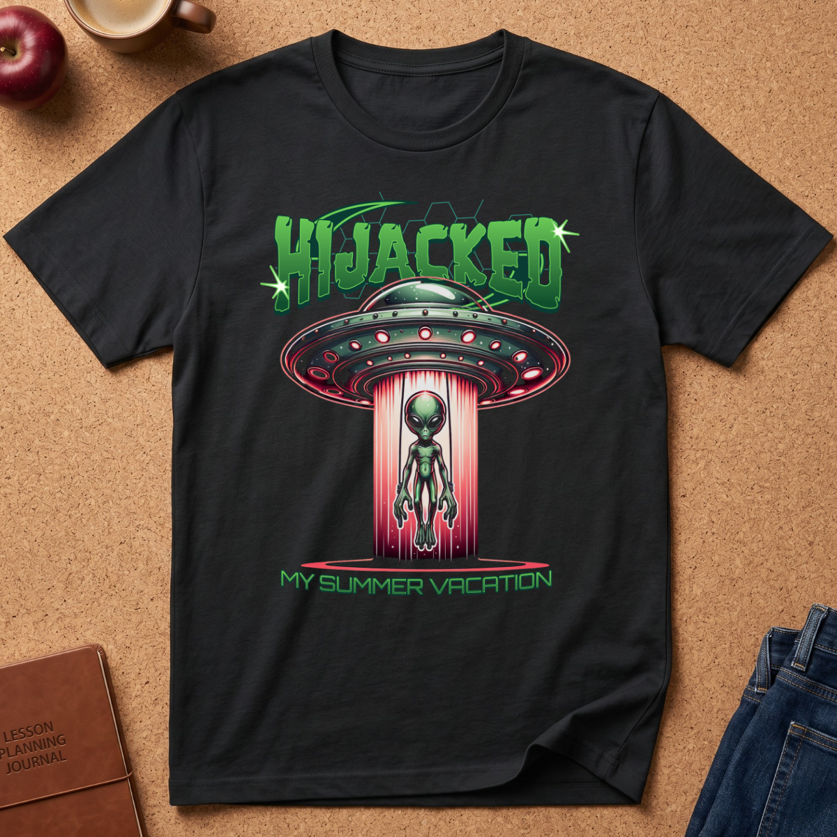 A black short-sleeve graphic T-shirt laid flat on a cork surface, featuring a green and pink retro UFO design with the text 'HIJACKED' and 'MY SUMMER VACATION.' Partial props around the shirt include an apple, a mug, a journal, and jeans.