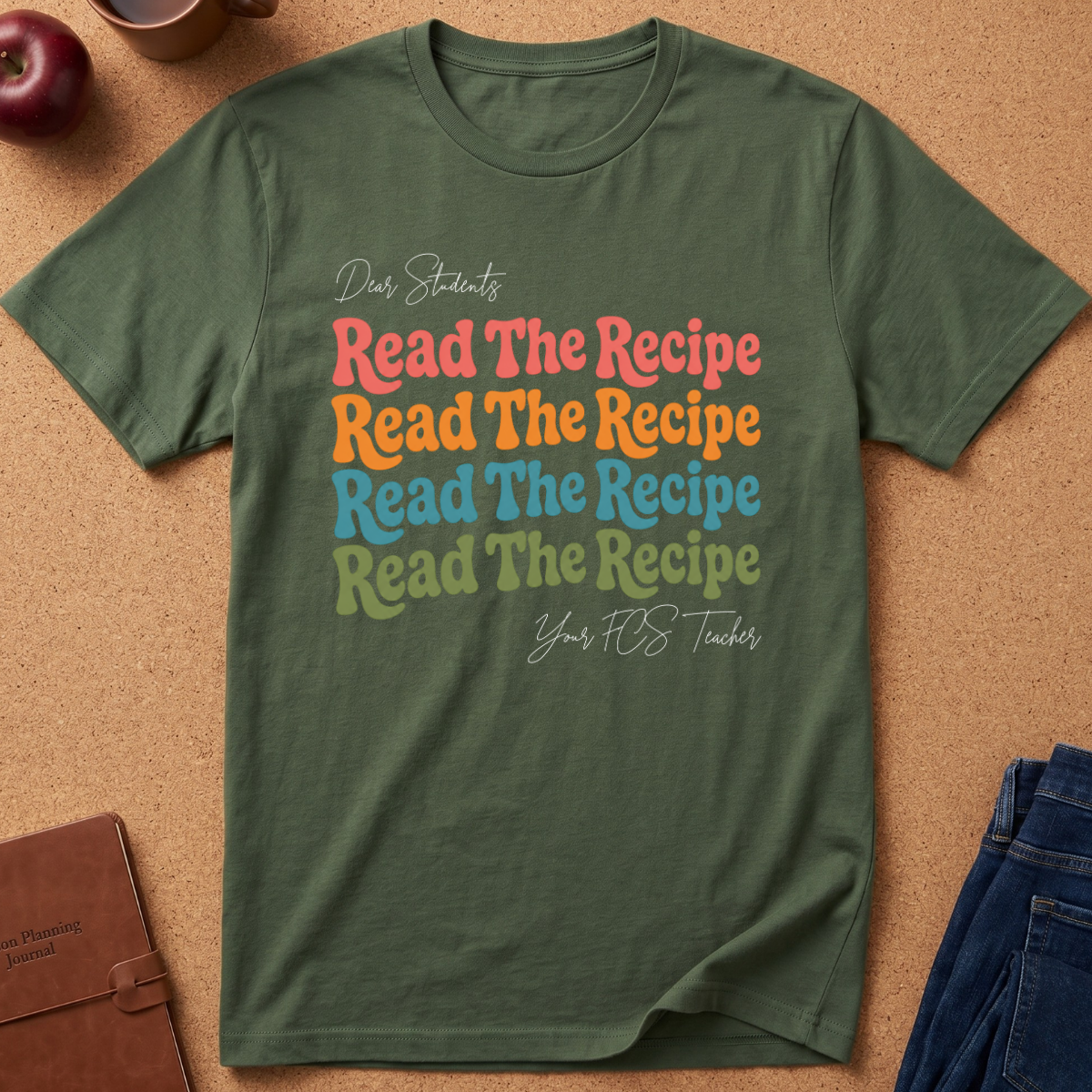 An olive green graphic t-shirt laid flat on a cork background, featuring a colorful retro-style print that reads 'Dear Students Read The Recipe Your FOS Teacher' repeated. Styled flat-lay props include a brown leather notebook, a red apple, and blue jeans.