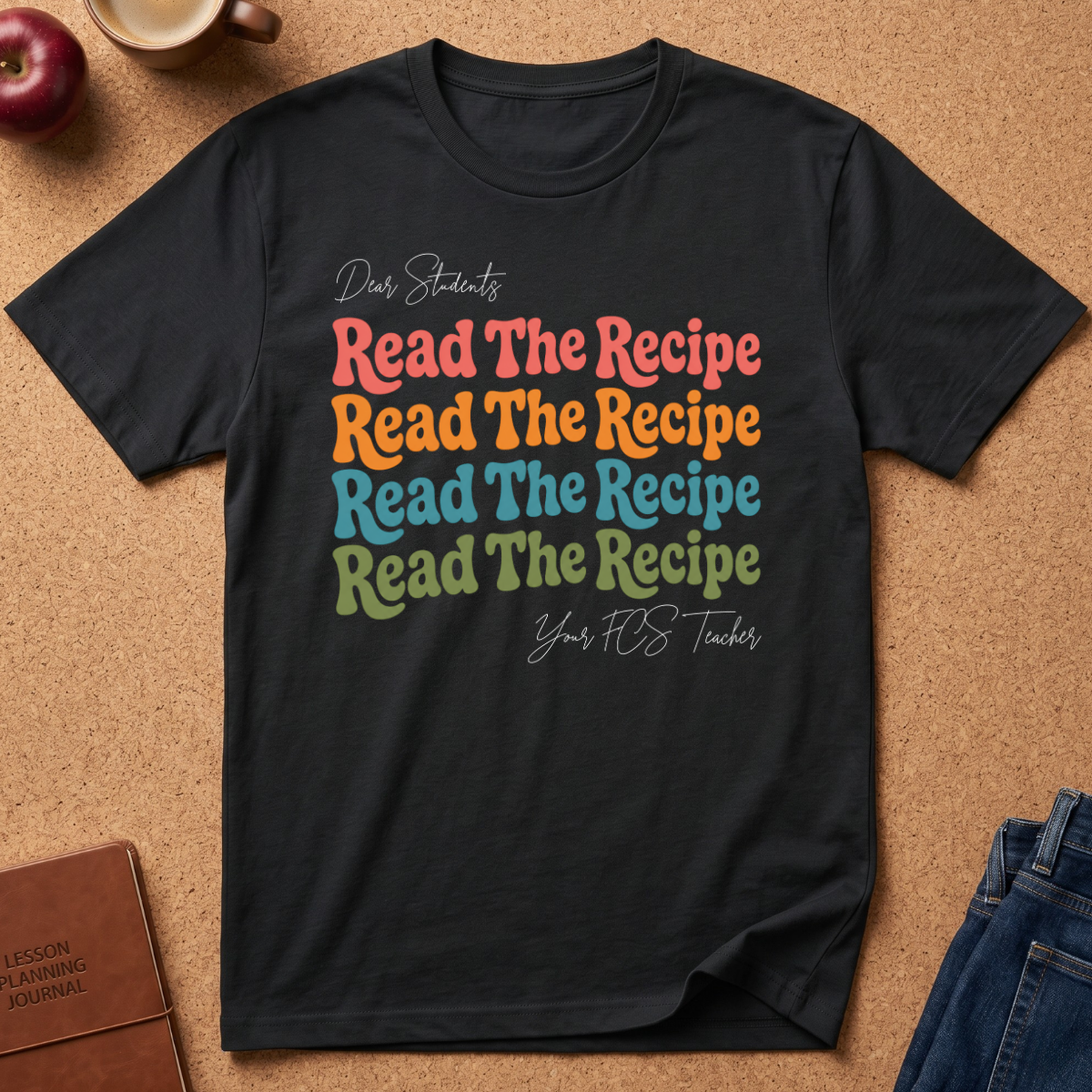A black graphic t-shirt laid flat on a cork background, featuring the text 'Dear Students Read The Recipe' repeated in pastel pink, orange, blue, and green, with 'Your FOS Teacher' in white script below.