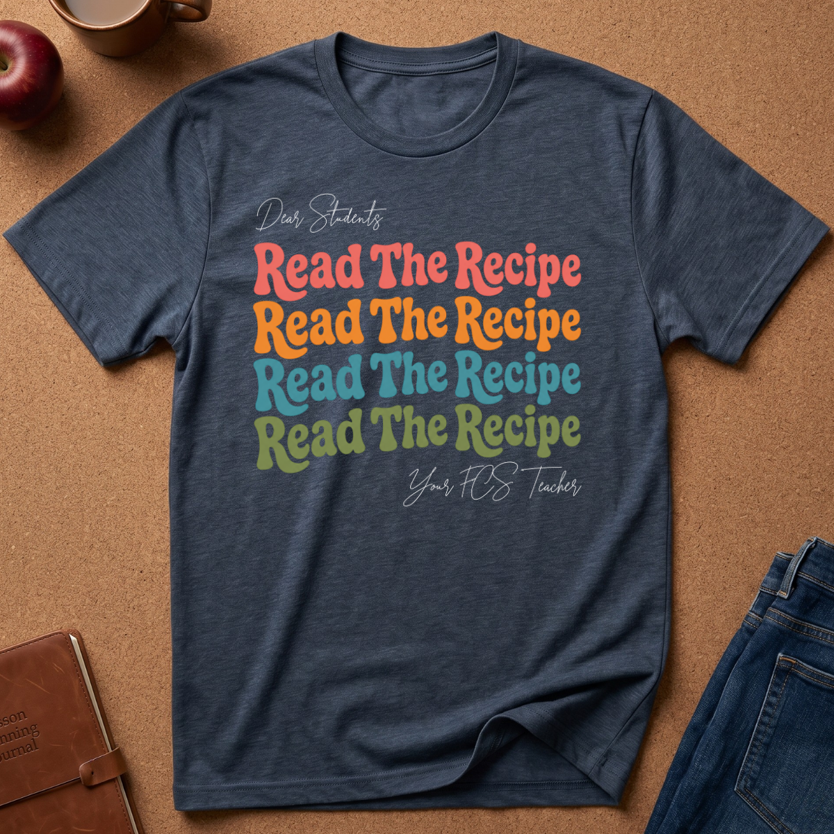 A navy blue heathered graphic t-shirt laid flat on a brown cork board background, surrounded by lifestyle props including a brown leather notebook, a red apple, a coffee mug, and blue jeans. The t-shirt features a colorful text graphic that reads 'Dear Students Read The Recipe Read The Recipe Read The Recipe Read The Recipe Your F*CS Teacher' in coral, orange, light blue, and olive green lettering.