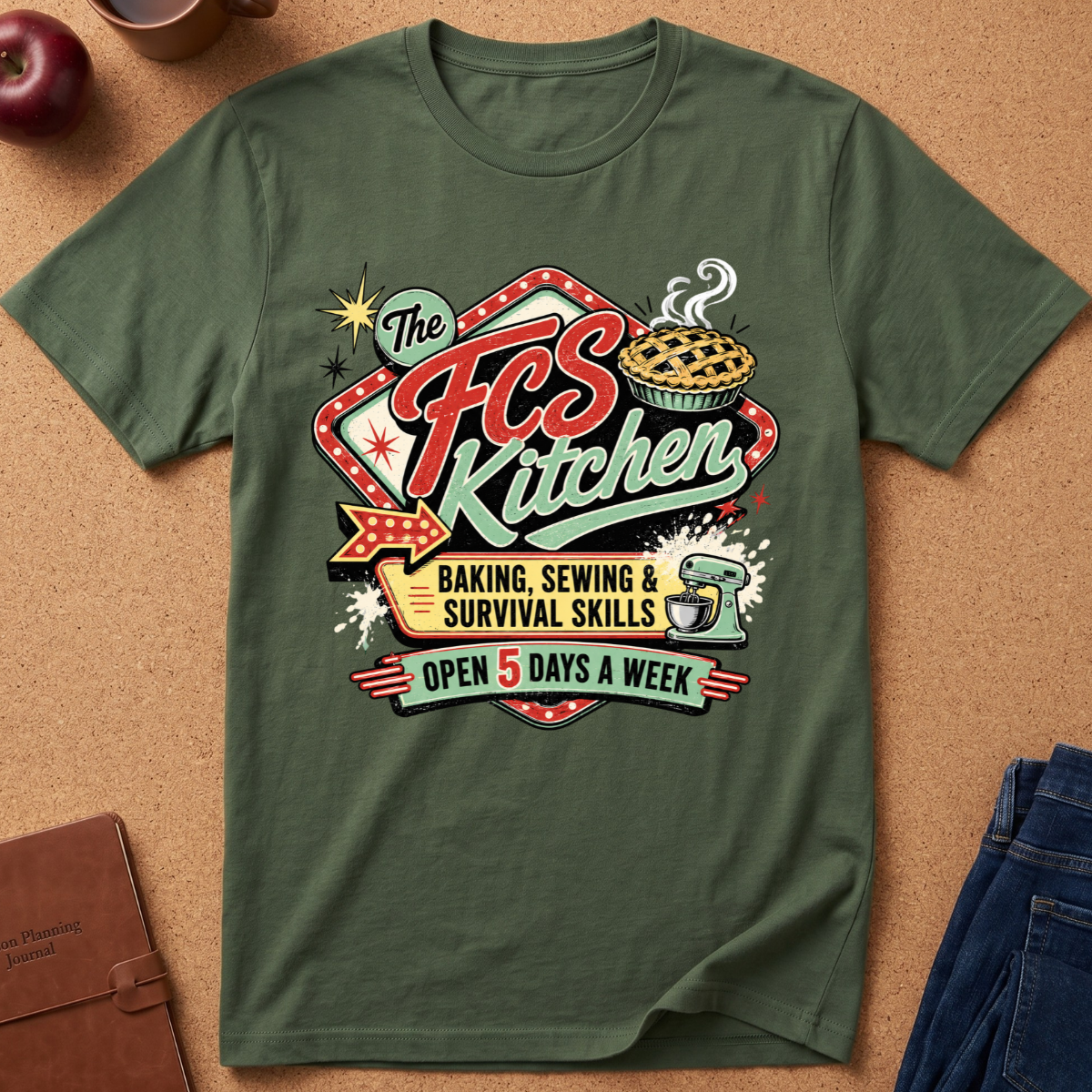 An olive green graphic t-shirt laid flat on a cork background, featuring a retro-style 'The FCS Kitchen' logo with illustrations of a pie, stars, and text reading 'Baking, Sewing & Survival Skills' and 'Open 5 Days A Week'.