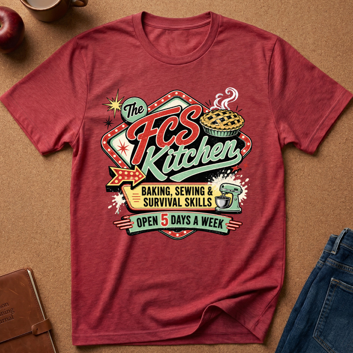 A heathered red graphic t-shirt laid flat on a brown textured surface, surrounded by lifestyle props including a coffee mug, an apple, a notebook, and blue jeans. The shirt features a retro-style 'The FCS Kitchen' graphic with baking, sewing, and survival skills text.