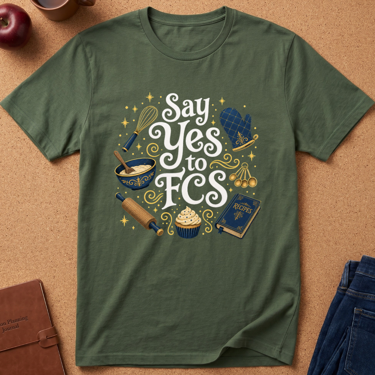An olive green short-sleeve t-shirt laid flat on a cork background, featuring a whimsical baking-themed graphic that reads 'Say Yes to FCS' in white script, surrounded by illustrations of a whisk, mixing bowl, cupcake, rolling pin, book, and cherries.