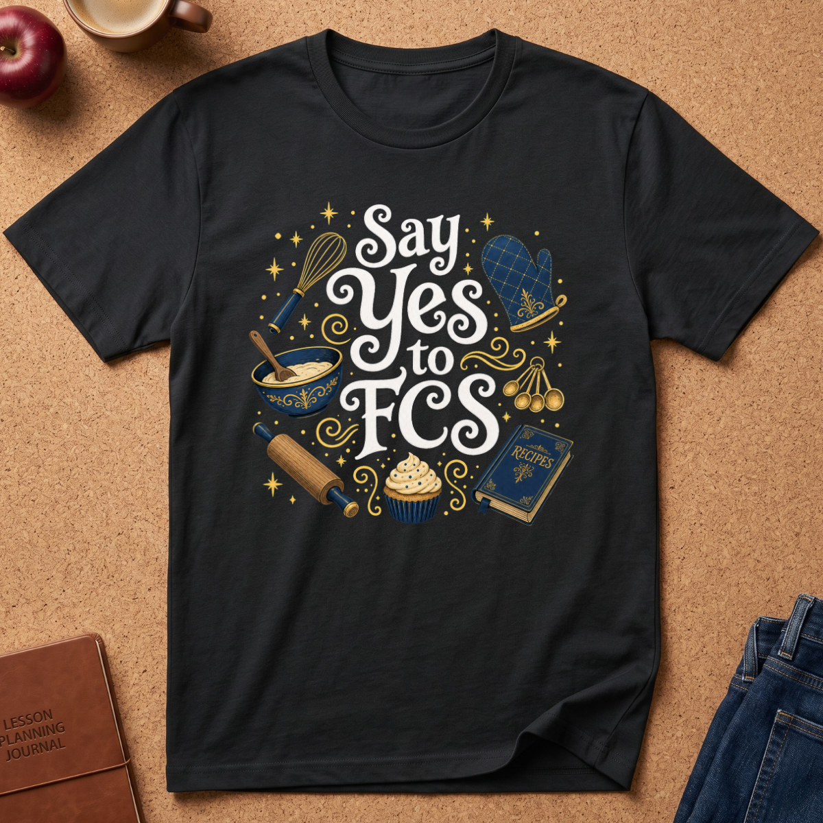 A black short-sleeve graphic T-shirt laid flat on a cork surface, featuring decorative text and baking-themed illustrations including a whisk, bowl, cupcake, rolling pin, oven mitt, and notebook. The shirt is styled with a notebook, apple, mug, and jeans around it.