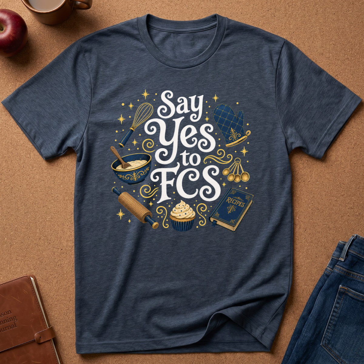 A navy blue heather graphic t-shirt laid flat on a brown cork background, surrounded by lifestyle props including an apple, a coffee mug, a leather journal, and folded blue jeans. The t-shirt features a whimsical graphic with the text 'Say Yes to FCS' in white and gold, surrounded by baking and academic motifs like a whisk, mixing bowl, rolling pin, cupcakes, and a gavel.