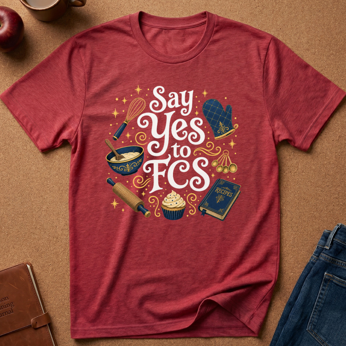 A heathered red graphic t-shirt laid flat on a brown cork background with styled props including an apple, a mug, a notebook, and jeans. The t-shirt features a 'Say Yes to FCS' graphic with baking and religious-themed illustrations like a whisk, mixing bowl, rolling pin, cupcakes, and a book.