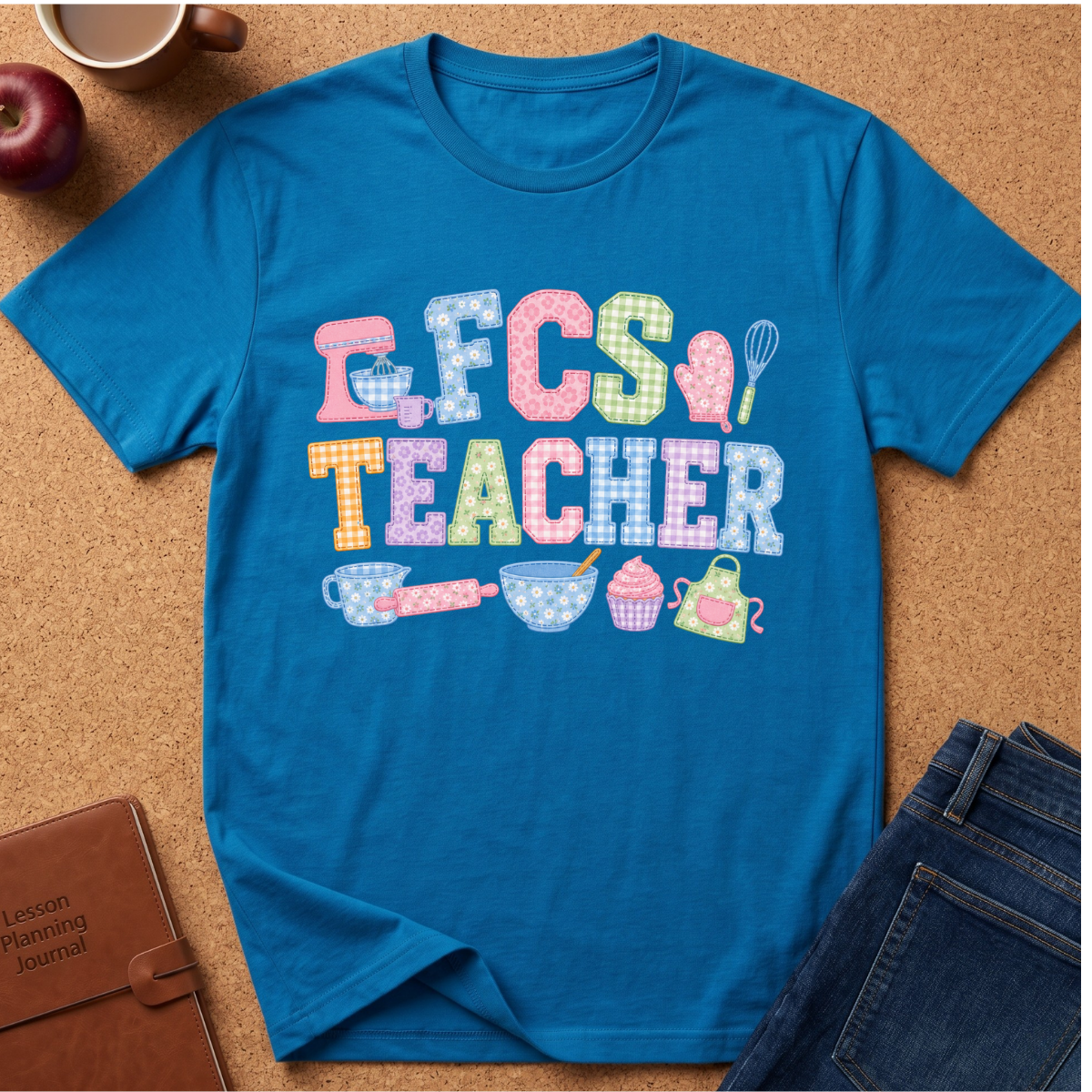 A bright blue graphic T-shirt laid flat on a cork surface, surrounded by styled props including an apple, a mug, a notebook, and jeans. The shirt features a colorful 'FCS TEACHER' design with playful kitchen-themed illustrations such as a stand mixer, spatula, whisk, teapot, cupcake, and baking pans.