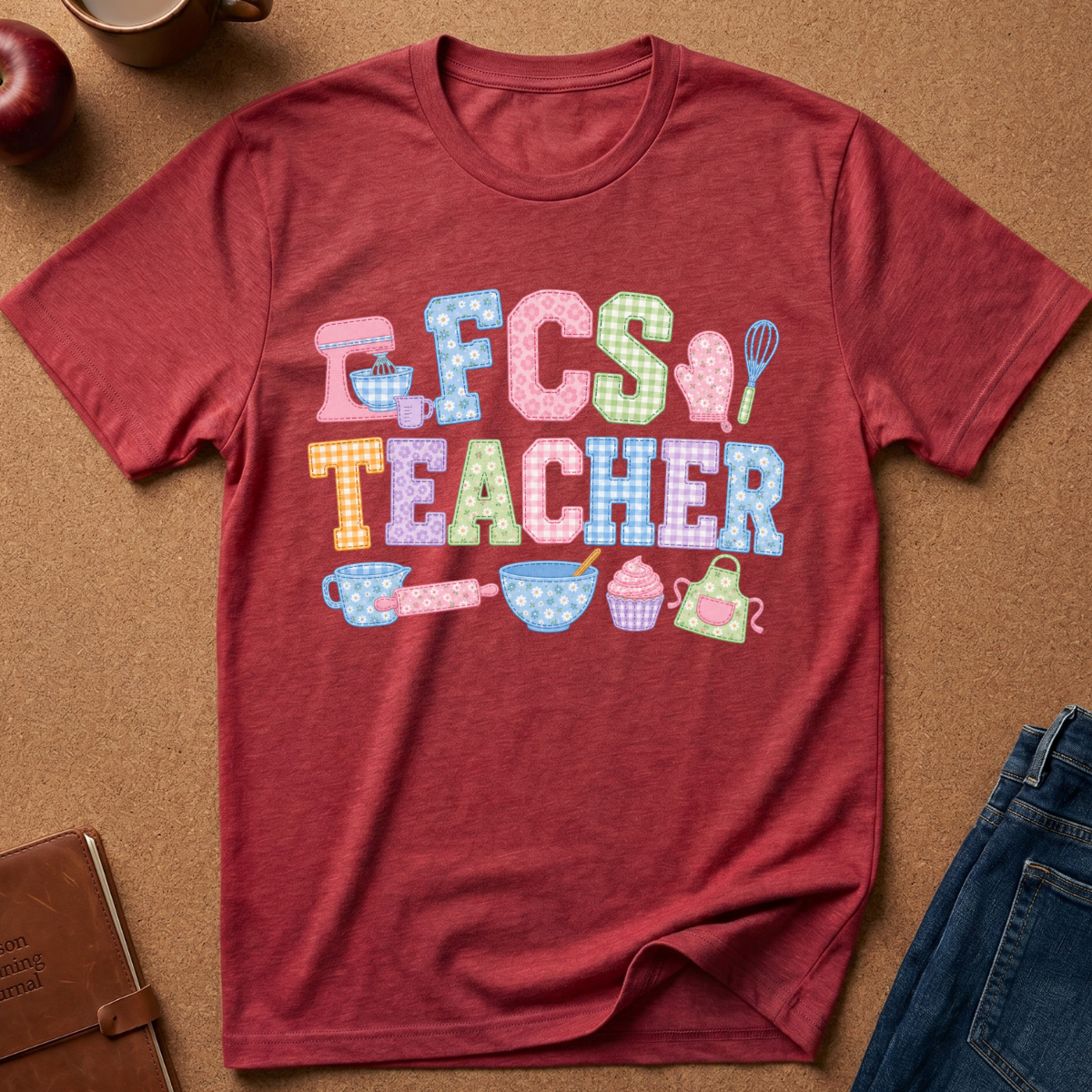 A heathered red short-sleeve t-shirt laid flat on a brown cork background, featuring a colorful 'FCS TEACHER' graphic with pastel gingham-style lettering and school-themed illustrations. Styled flat-lay props include an apple, coffee cup, notebook, and blue jeans around the shirt.