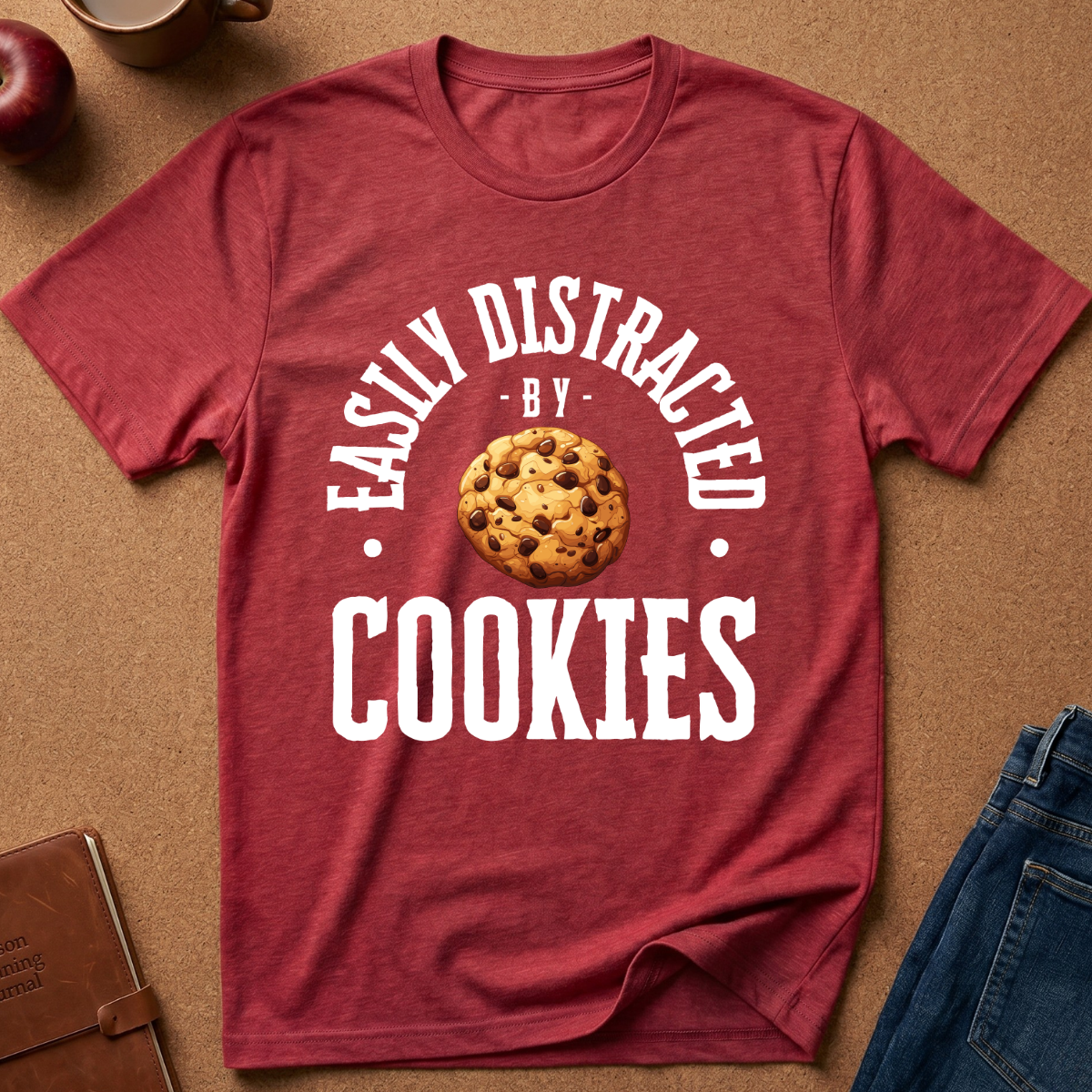 A heathered red short-sleeve t-shirt laid flat on a brown textured background, featuring a white graphic that reads 'EASILY DISTRACTED BY COOKIES' around an illustration of a chocolate chip cookie. Styled flat-lay props include a brown notebook, a red apple, a coffee cup, and blue jeans.