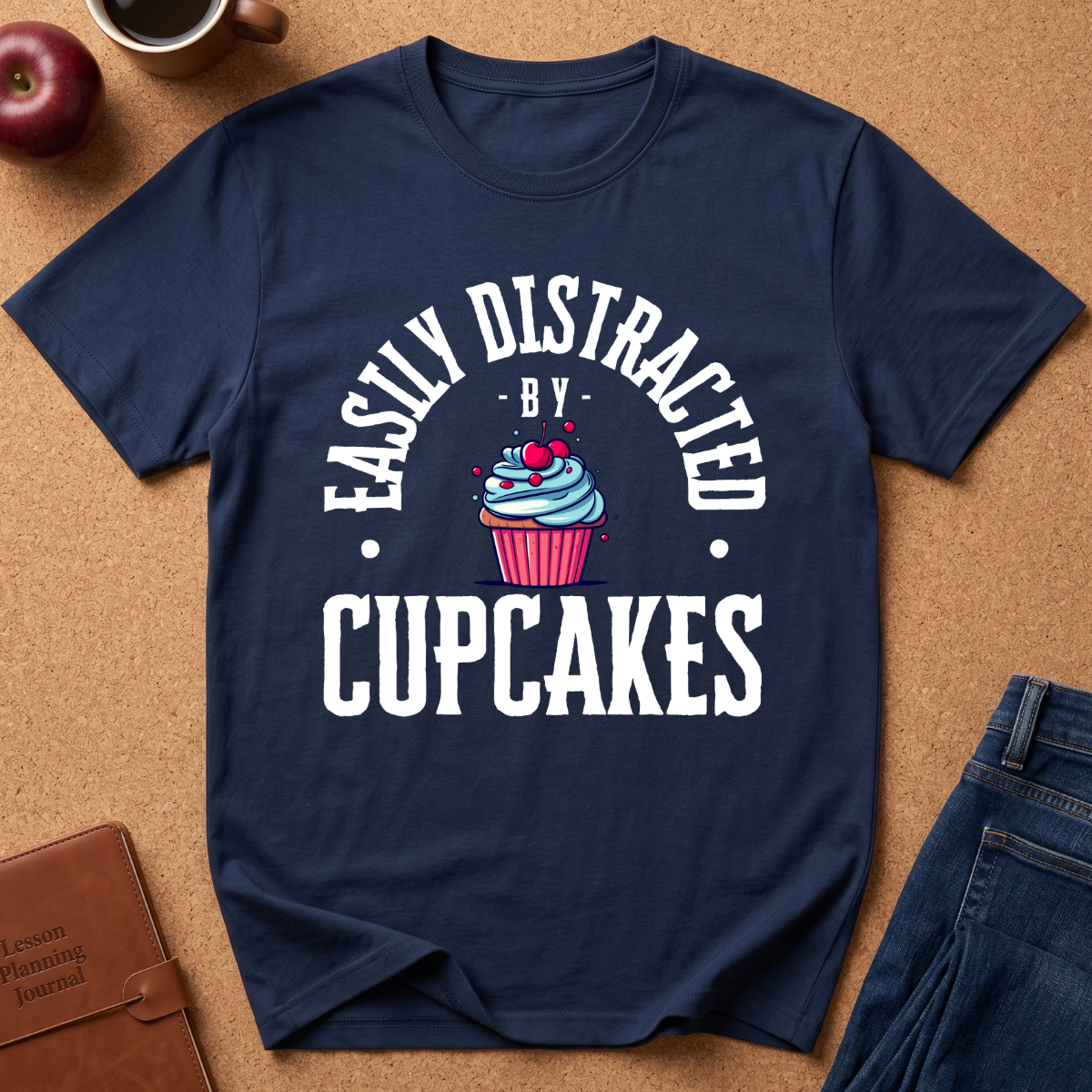 A navy blue graphic t-shirt laid flat on a brown cork background, featuring the white text 'EASILY DISTRACTED BY CUPCAKES' and a cupcake illustration in the center. A red apple, a coffee cup, a brown journal, and blue jeans are styled around the shirt.