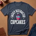 A navy blue heather graphic t-shirt laid flat on a brown cork background, surrounded by lifestyle props including an apple, a coffee mug, a notebook, and folded blue jeans. The t-shirt features a white arched text graphic reading 'EASILY DISTRACTED BY CUPCAKES' surrounding an illustration of a cupcake with blue frosting, a pink wrapper, and a red cherry.