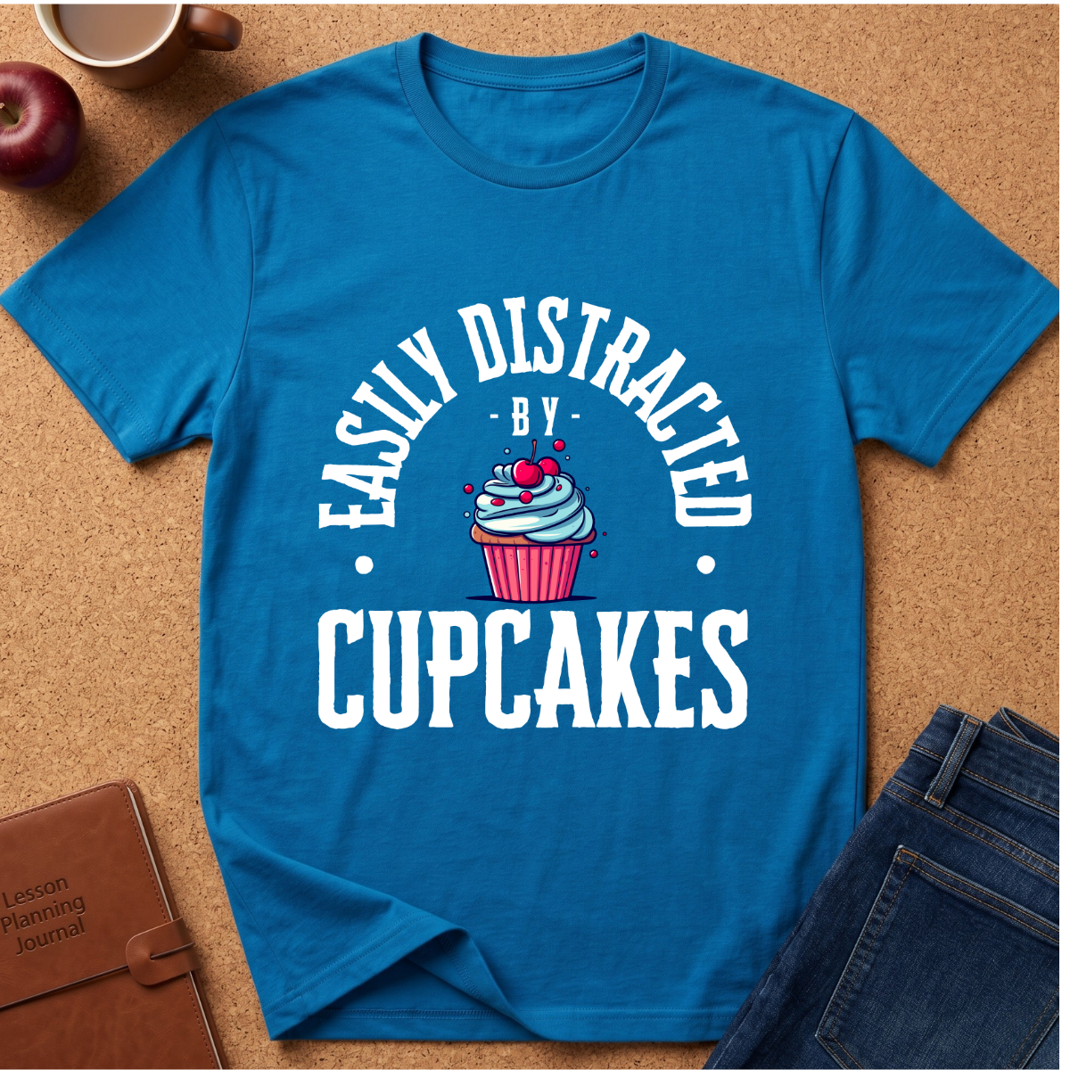 A bright blue short-sleeve graphic T-shirt laid flat on a cork background, featuring the phrase 'EASILY DISTRACTED BY CUPCAKES' in white around an illustrated cupcake with pink frosting and red cherries. Surrounding props include a brown notebook, a red apple, a mug, and blue jeans.