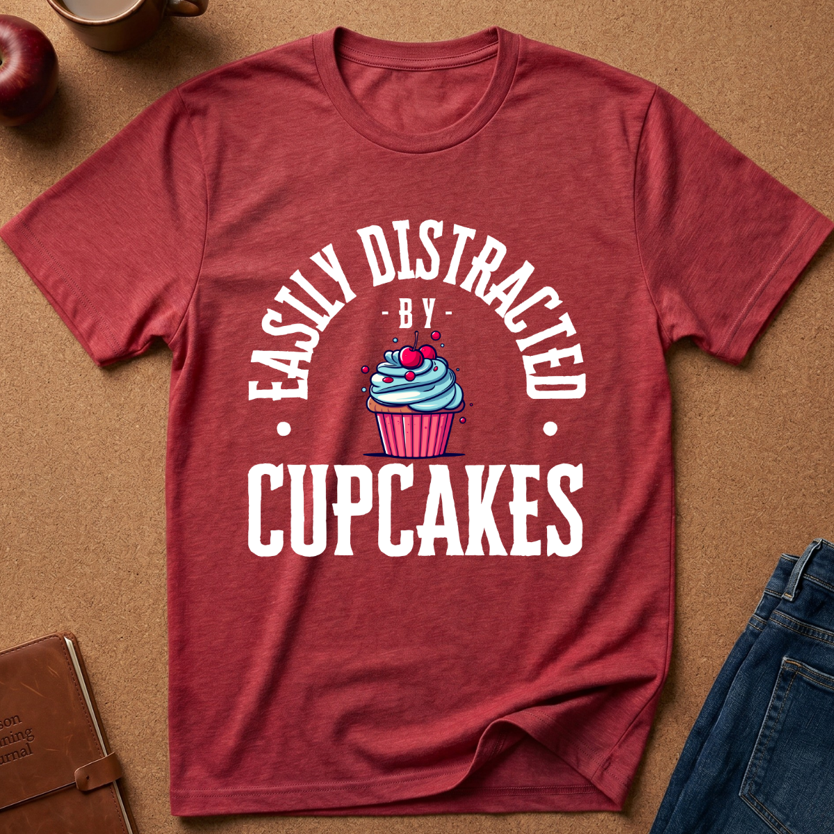 A heathered red short-sleeve t-shirt laid flat on a brown textured background, featuring a white graphic that reads 'EASILY DISTRACTED BY CUPCAKES' around a cupcake illustration with pink frosting and a cherry.